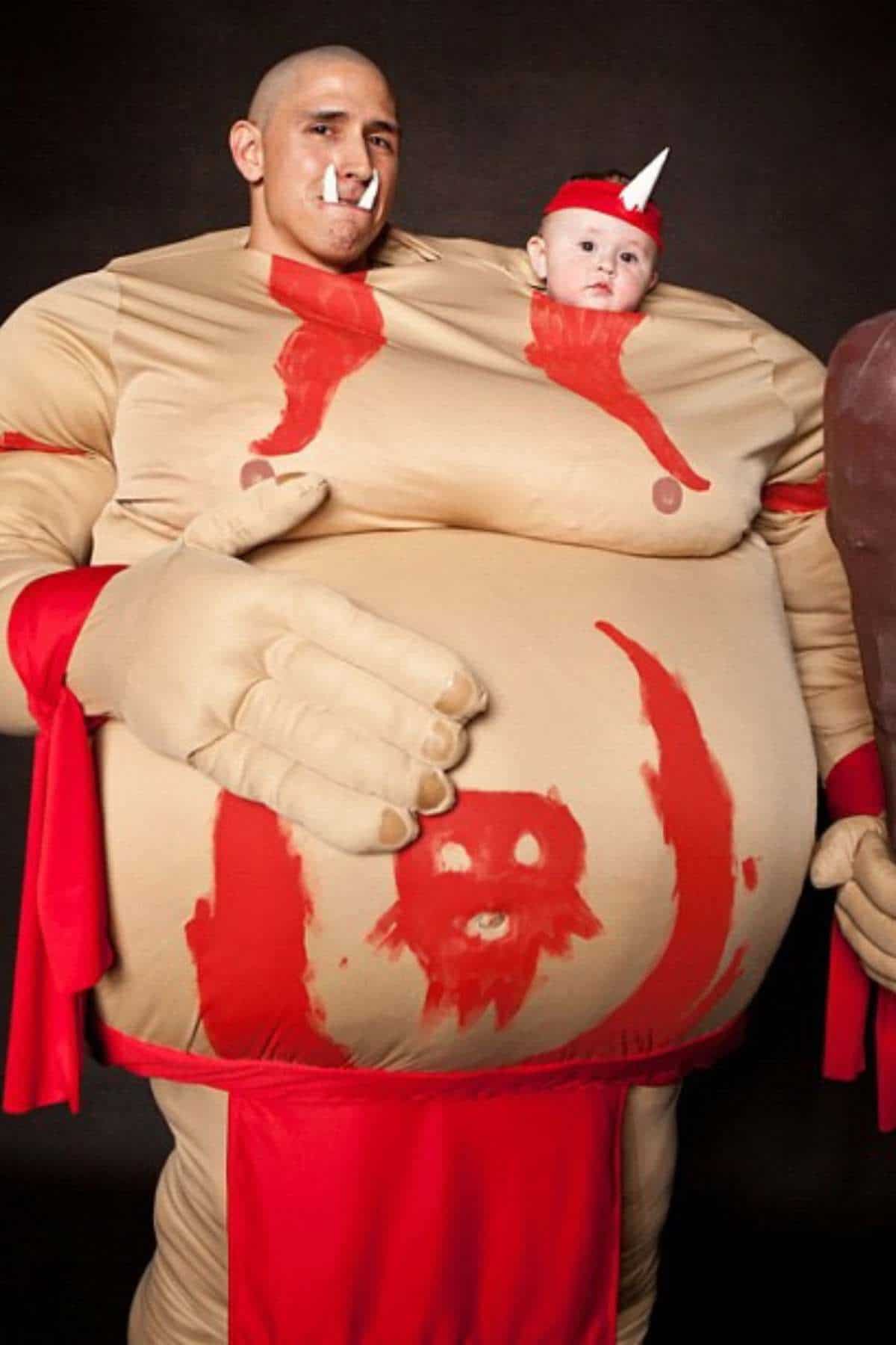 An adult in a muscular, painted costume with fake blood holds a baby in a headband; the baby appears to be attached to the costume's stomach.