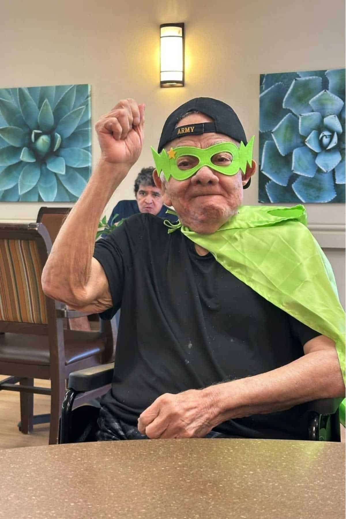 An older man wearing a green superhero mask and cape sits at a table, raising his hand. Another person is seated in the background.