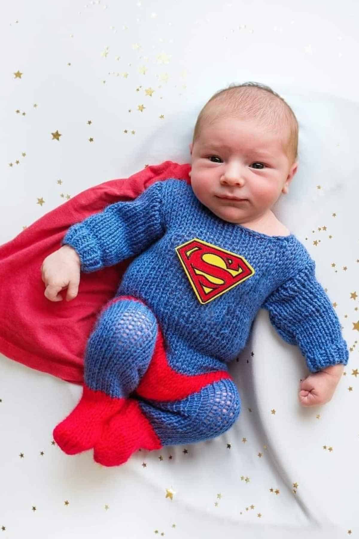 A baby dressed in a knitted blue outfit with a Superman logo, red cape, and red booties, lying on a white blanket decorated with gold stars.