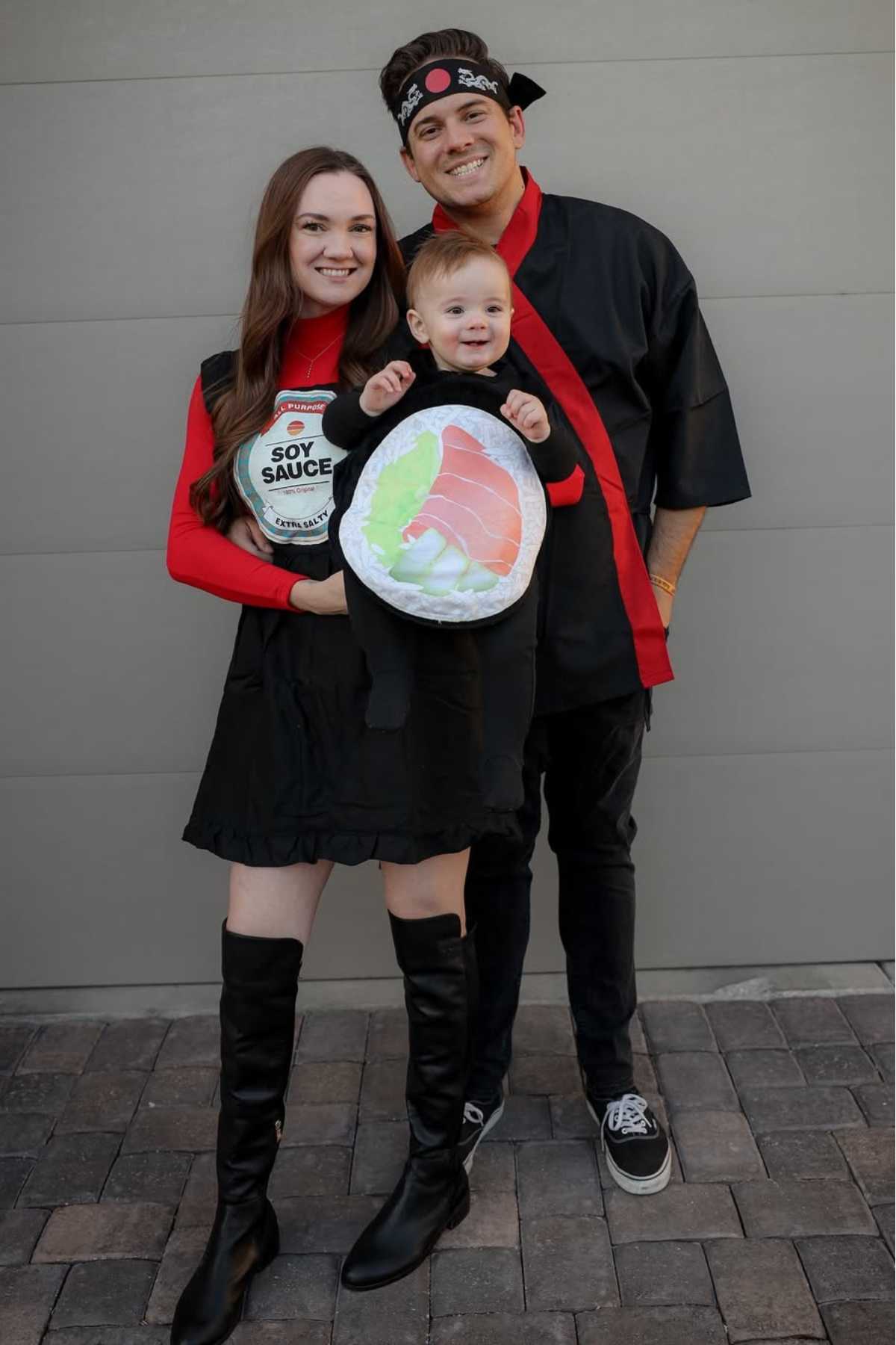 A smiling family in themed costumes: a woman dressed as soy sauce, a man in sushi chef attire, and a baby in a sushi roll costume. They stand together in front of a gray wall.