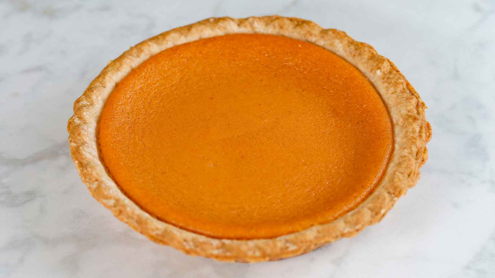 A whole pumpkin pie with a golden-brown crust and smooth orange filling, placed on a light marble surface.