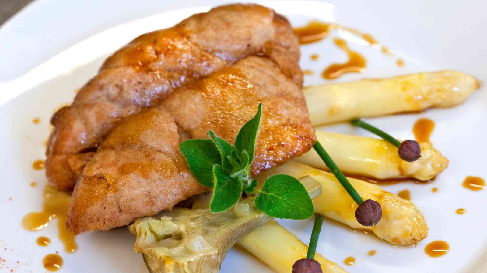Two pieces of cooked meat served on white asparagus spears, garnished with fresh herbs, chives, and a drizzle of brown sauce on a white plate.