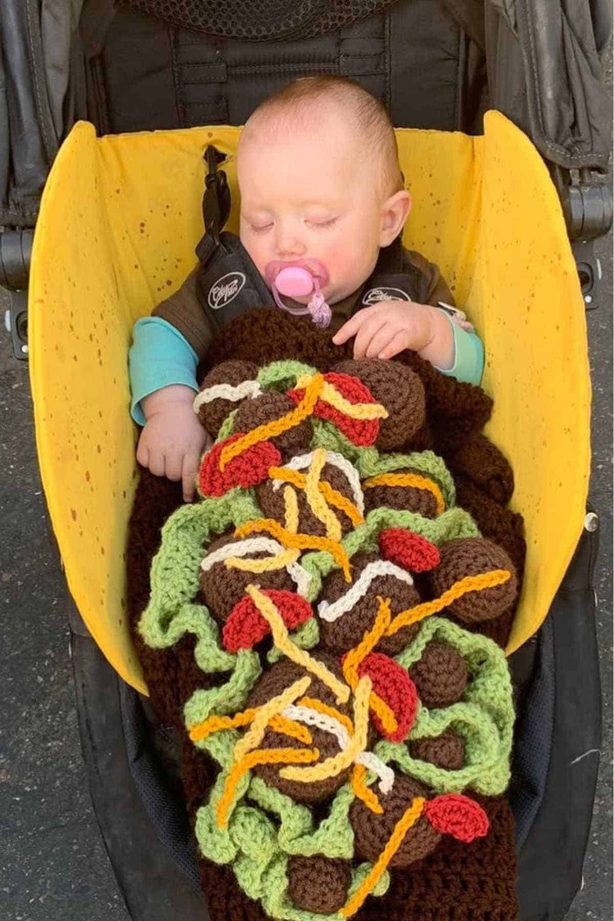 A baby sleeps in a stroller, covered with a crocheted blanket designed to look like a taco with lettuce, and other toppings.
