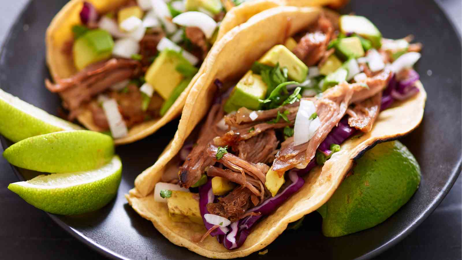 Two tacos filled with shredded meat, avocado, onions, cilantro, and cabbage on a black plate with lime wedges.