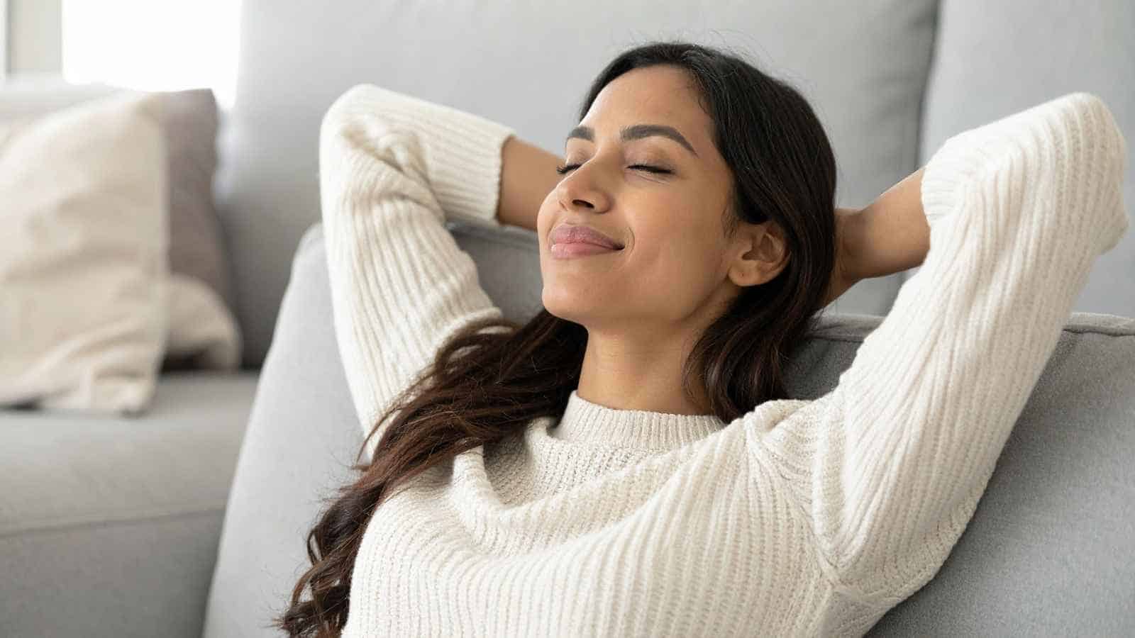 A woman in a white sweater relaxes on a couch with her hands behind her head, eyes closed, and a content expression.