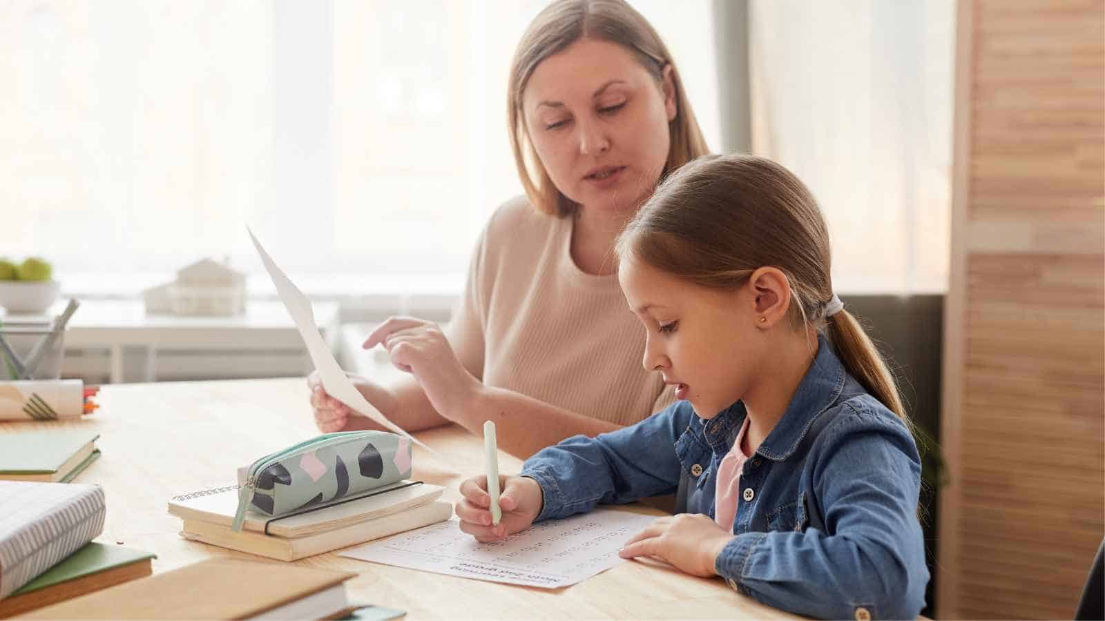 An adult helps a young girl with her homework at a desk. The girl is writing, and the adult is holding a sheet of paper.