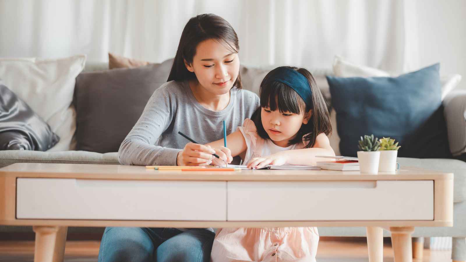 An adult and a child sit at a low table, drawing or writing on paper with colored pencils in a living room.