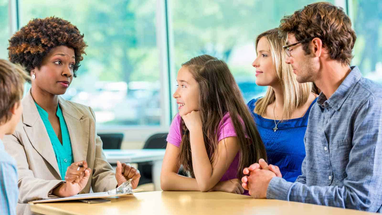 A woman in a blazer talks to a family of four at a table, possibly in a meeting or consultation, with windows and trees in the background.