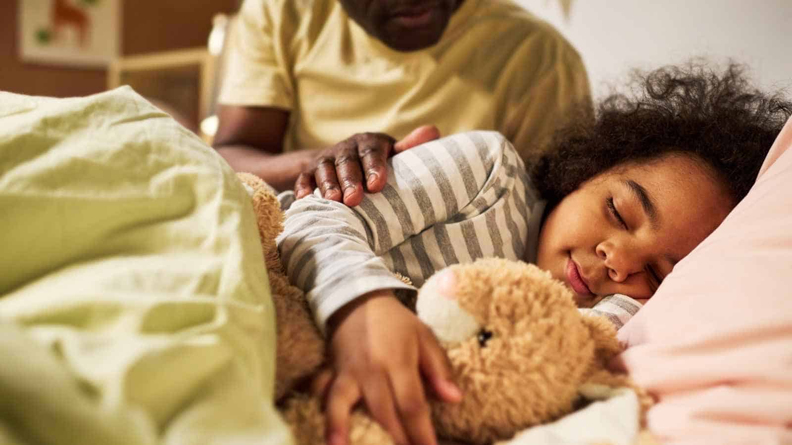 A child sleeps in bed hugging a stuffed animal while an adult gently rests a hand on the child’s back.