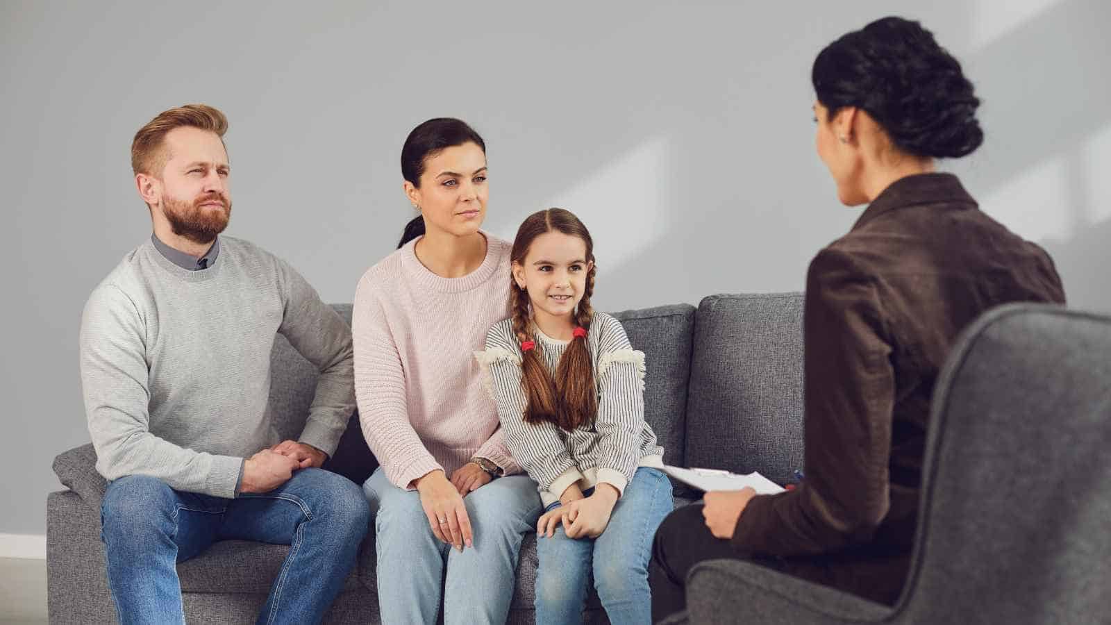 A man, woman, and young girl sit on a gray couch facing a woman with a notepad, suggesting a family counseling or therapy session in a modern office.