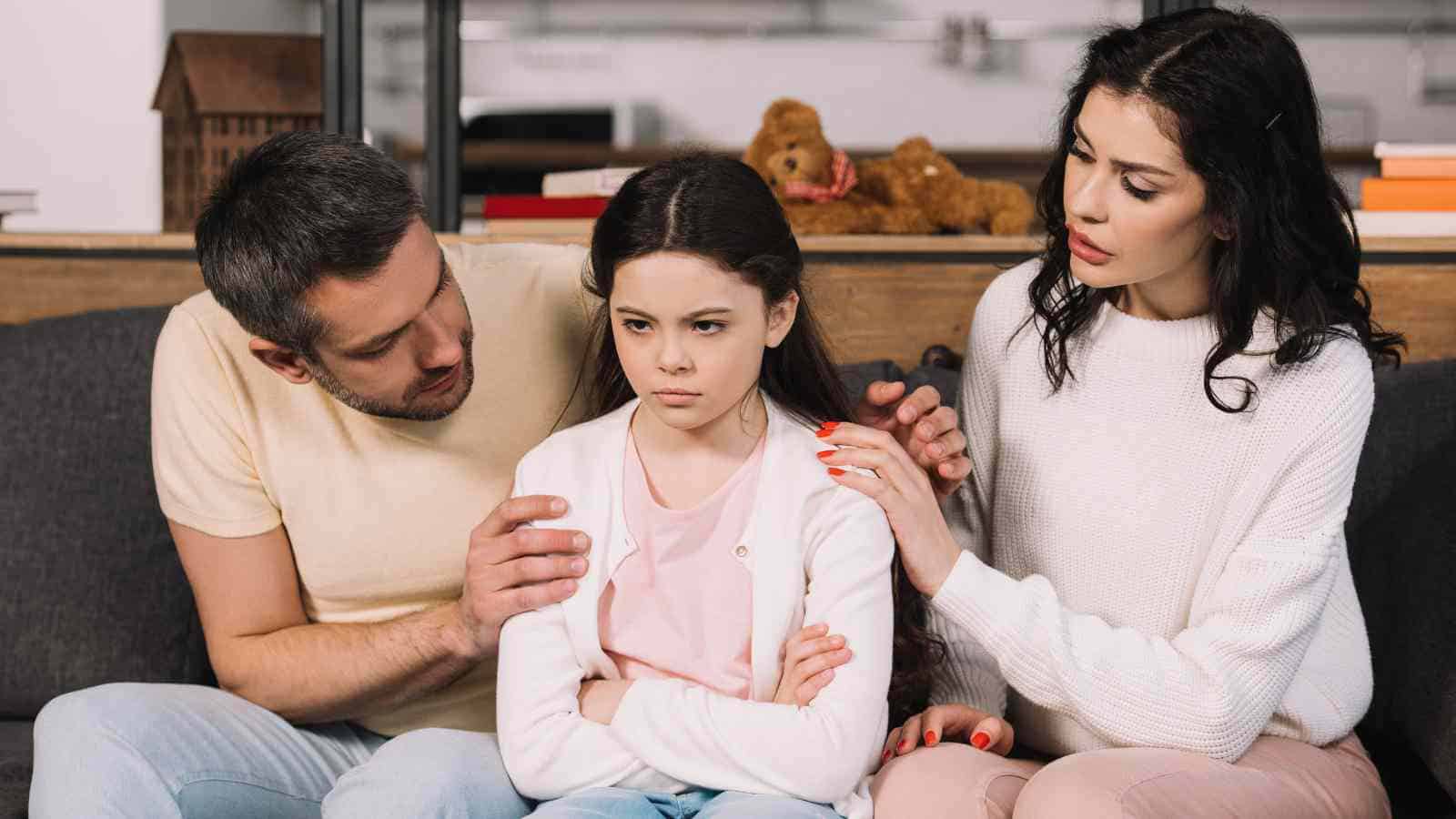 A young girl sits on a couch with her arms crossed, looking upset, while two adults sit beside her, appearing concerned and comforting.