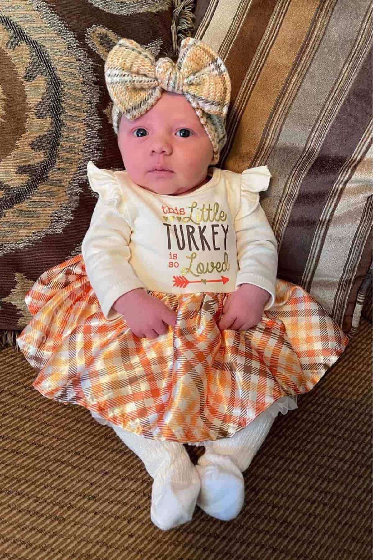 A baby wearing a plaid bow headband and a dress reading "This little turkey is so loved" sits on a patterned couch.