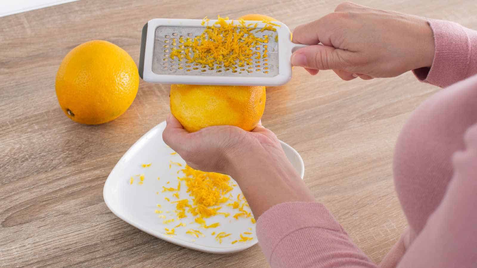 Person grating the zest from an orange over a white plate, with another whole orange on a wooden surface in the background.