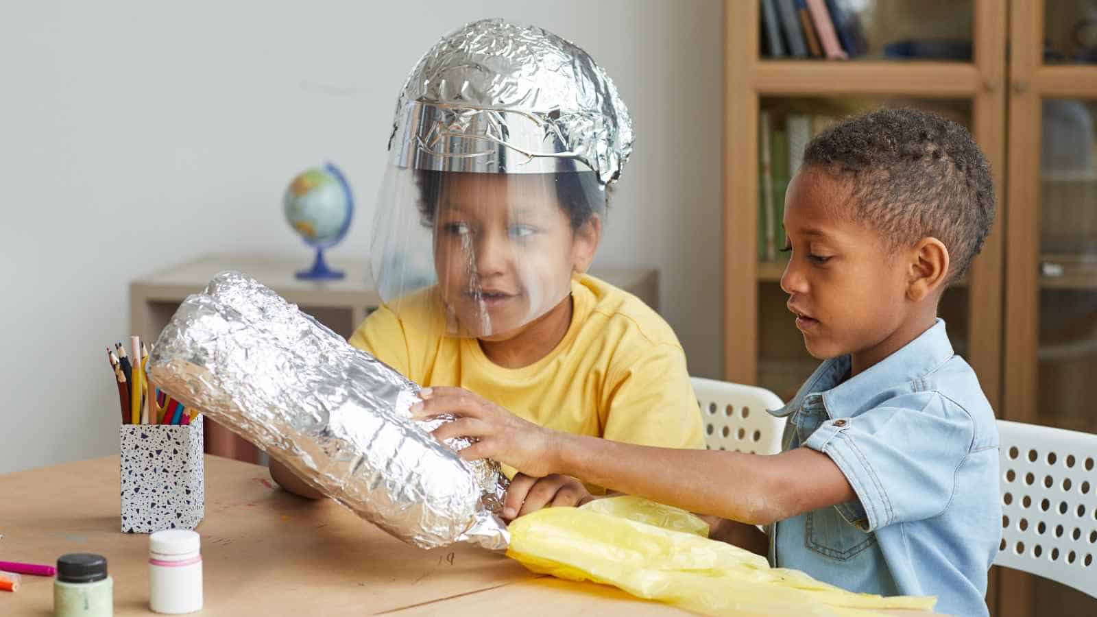 Two children sit at a table, one wearing a helmet and holding a large object made of aluminum foil, as the other child helps assemble it.