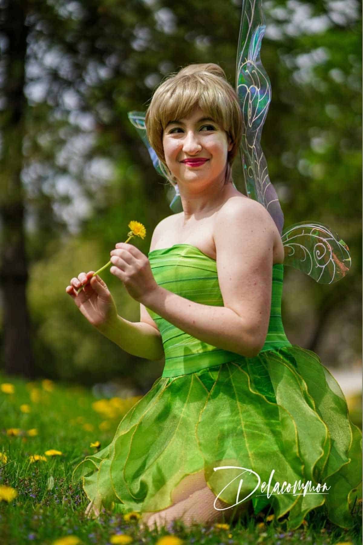 Person in a green fairy costume with wings sits on grass holding a yellow dandelion, smiling, with trees in the background.