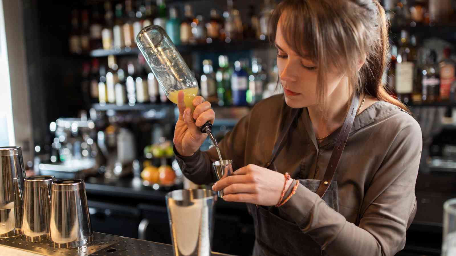 A bartender pours a yellow liquid from a bottle into a jigger at a bar with shelves of liquor bottles in the background.