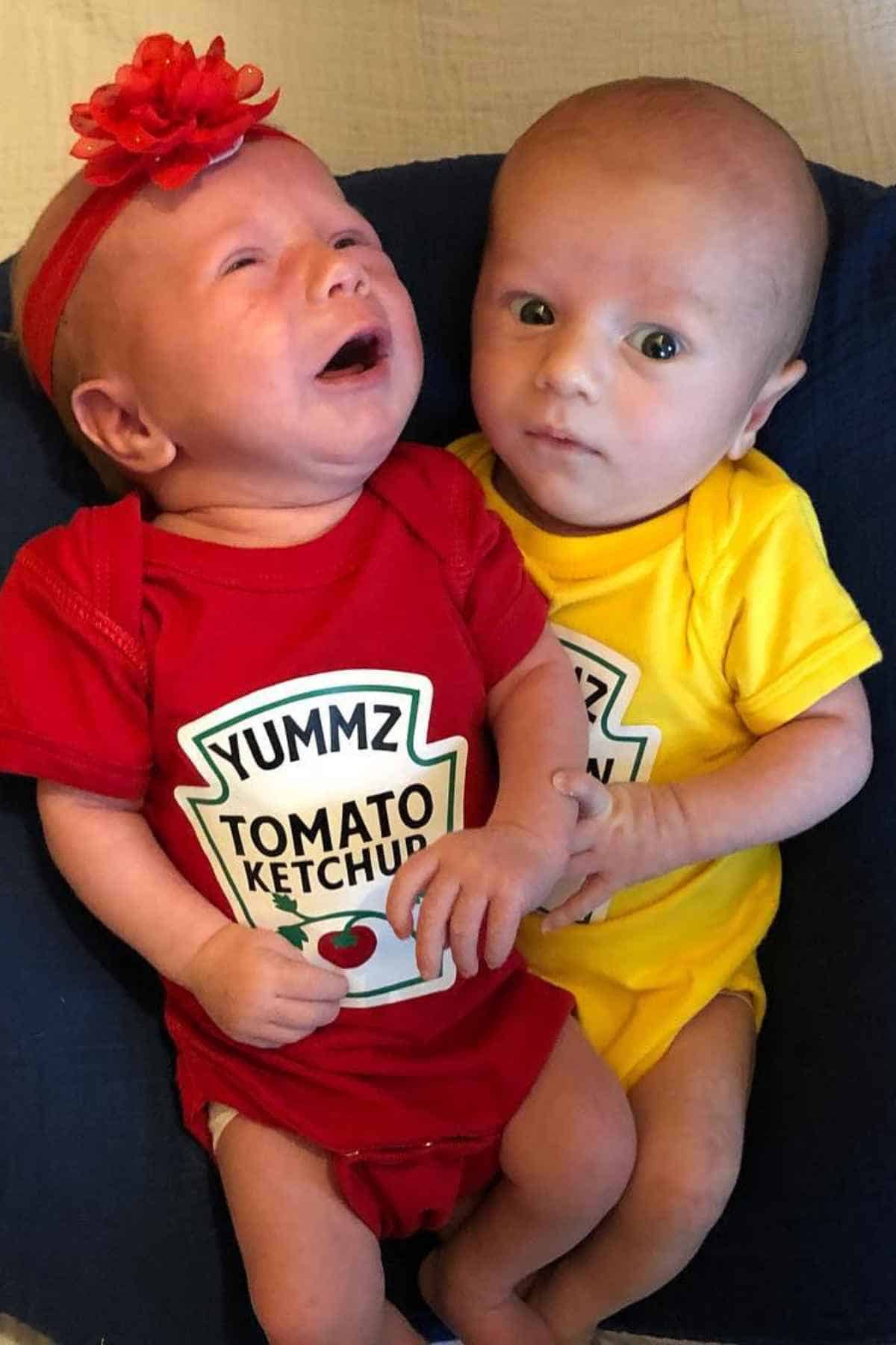 Two babies sit together; one wears a red "Yummz Tomato Ketchup" onesie and a red headband, while the other wears a yellow "Yummz" onesie. The baby in red appears to be crying.