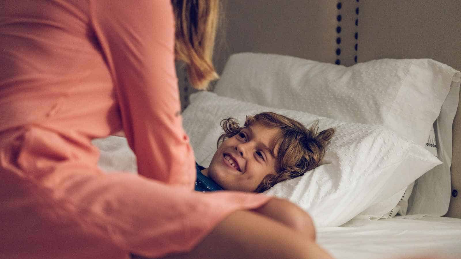 A smiling child lies on a bed with white pillows, looking up at an adult sitting beside them.