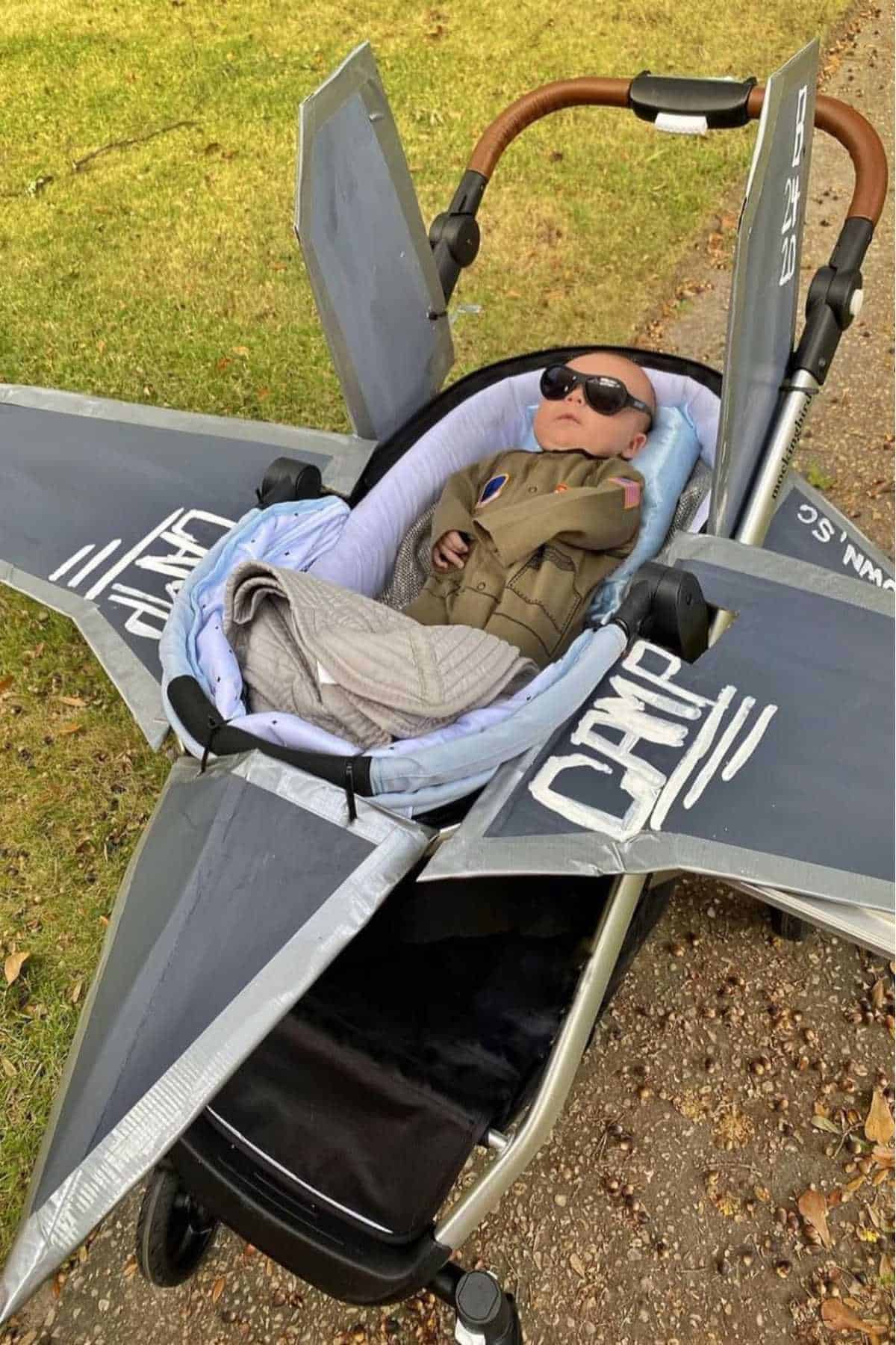 A baby wearing sunglasses and a jacket lies in a stroller decorated to look like a fighter jet, outdoors on a grassy path.