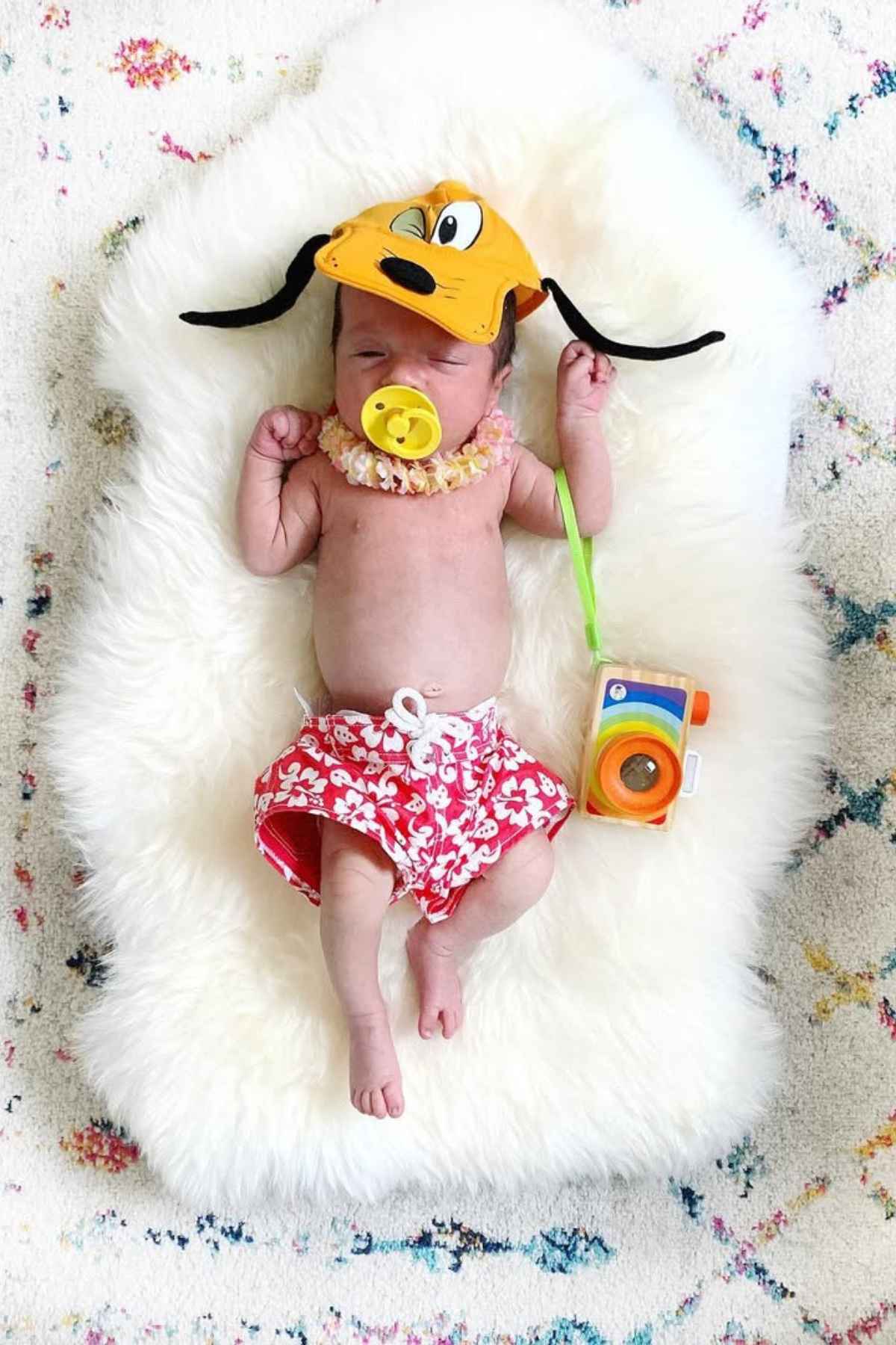 Baby wearing a Pluto hat, pacifier, floral shorts, and lei, holding a toy camera, lying on a fluffy white rug with a patterned background.