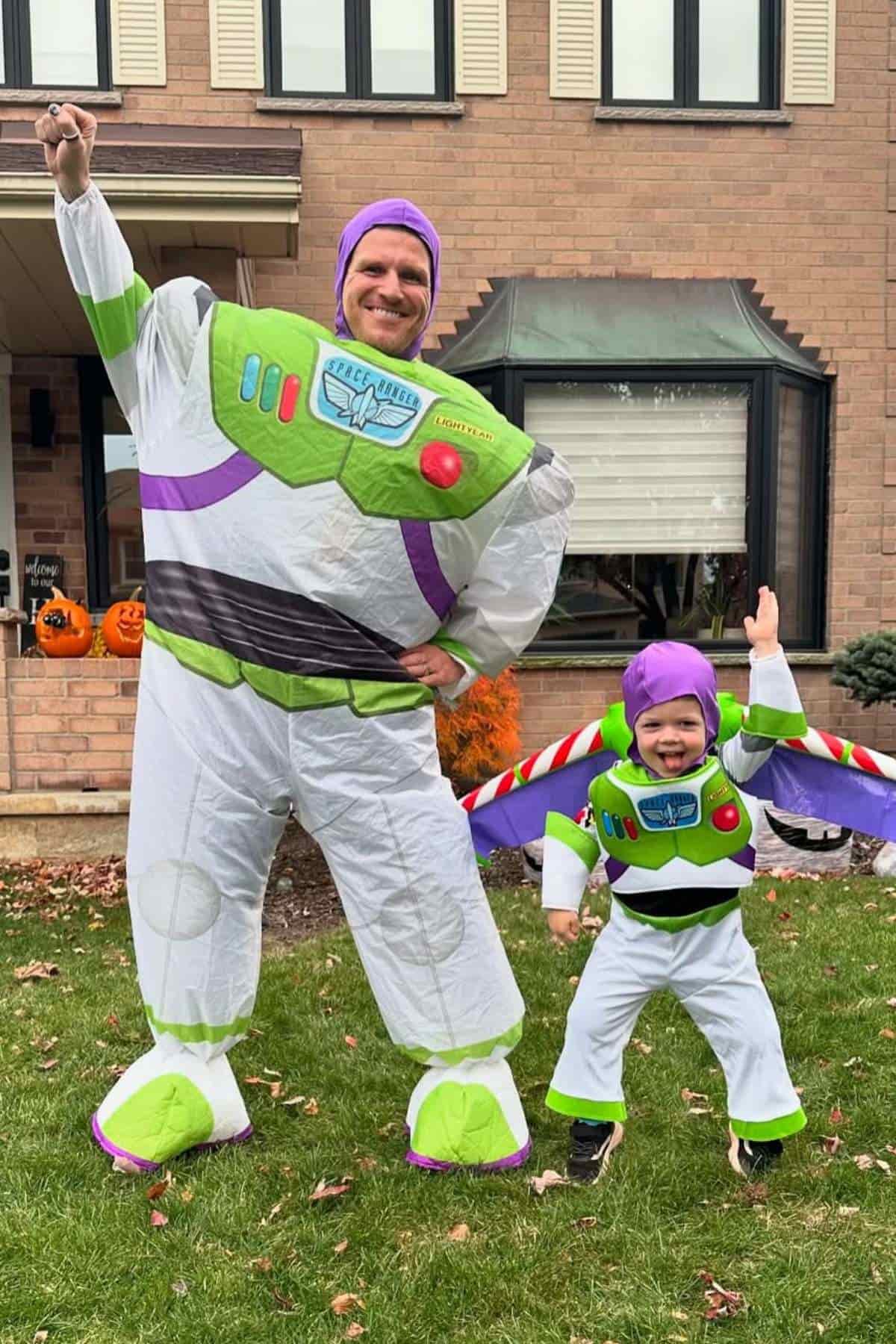 An adult and a child dressed in matching Buzz Lightyear costumes pose outside in front of a house, both with one arm raised.