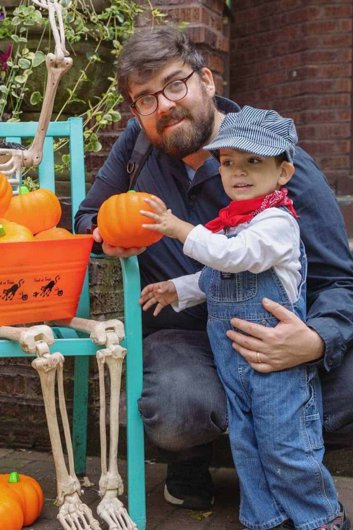An adult and a child dressed as a train conductor pose together outdoors near a skeleton decoration and pumpkins during Halloween.