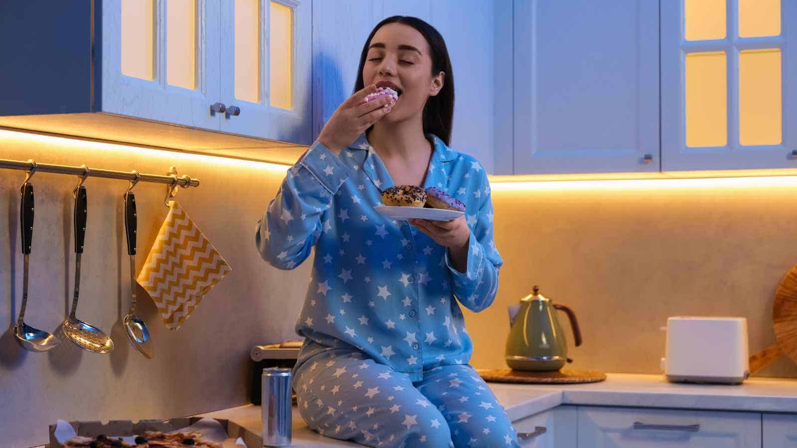 Woman in blue star-patterned pajamas sits on a kitchen counter, eating a donut with closed eyes and holding a plate, surrounded by kitchen appliances and utensils.