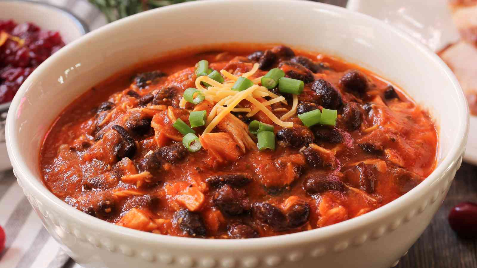 A white bowl filled with chili containing black beans, tomatoes, and meat, topped with shredded cheese and chopped green onions.