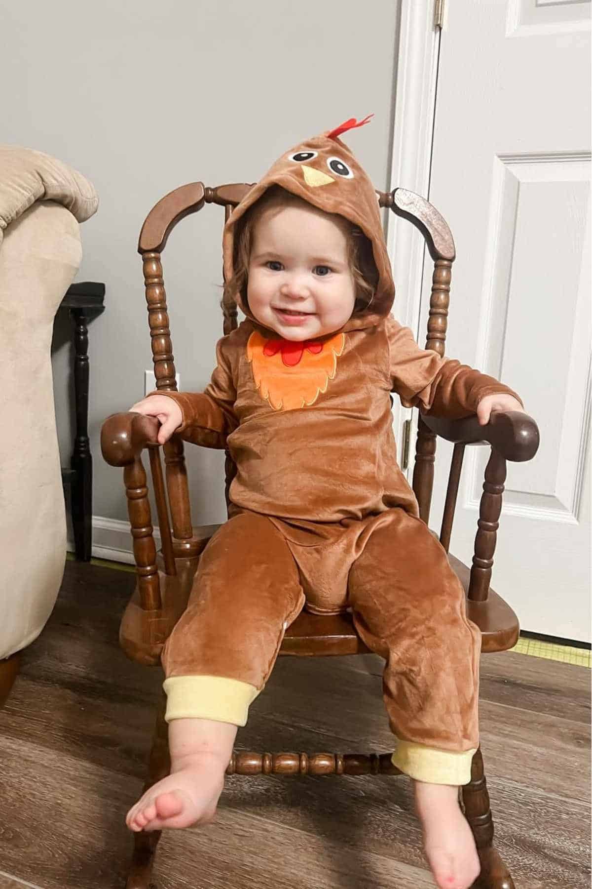 A baby dressed in a brown turkey costume with an orange beak sits and smiles on a wooden rocking chair indoors.