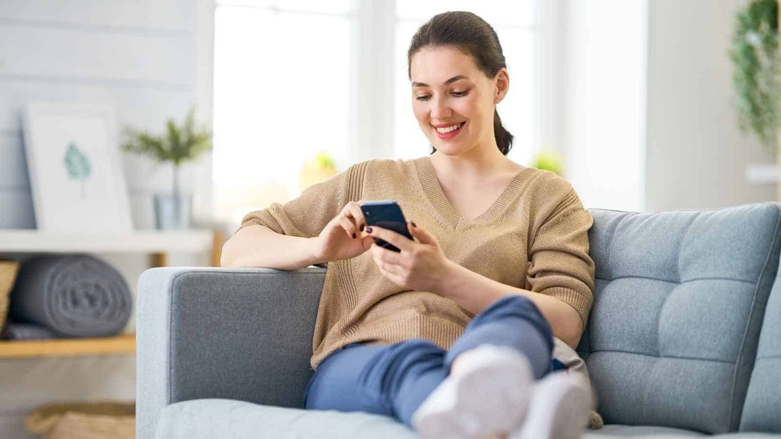 A woman sits on a sofa, smiling while using a smartphone. She is wearing a beige sweater and blue pants in a brightly lit living room.