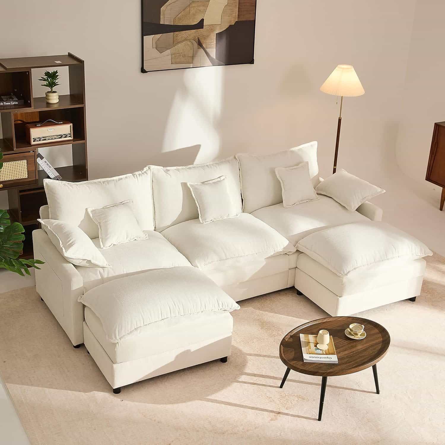 A white sectional sofa with multiple cushions sits in a bright living room, accompanied by a round wooden coffee table, floor lamp, bookshelf, and abstract wall art.