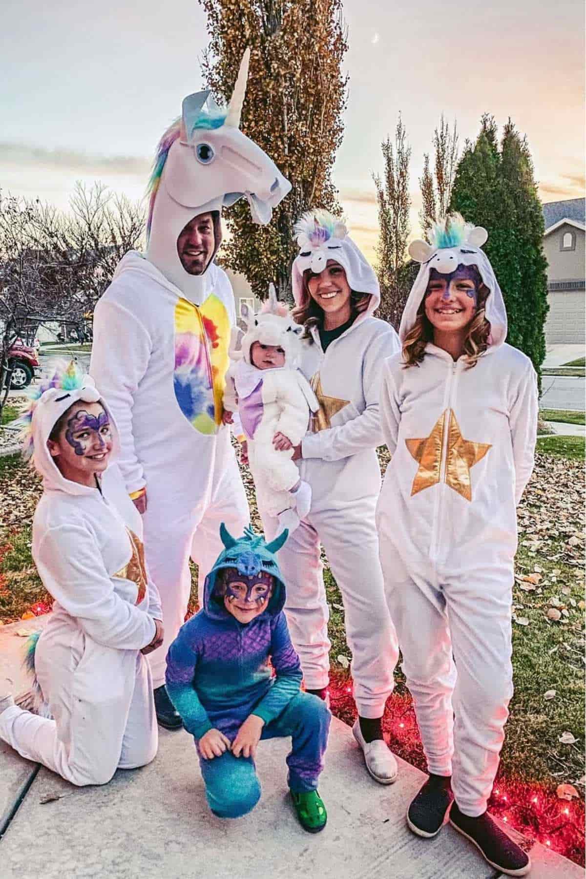 Six people, including one baby, are dressed in unicorn and dragon costumes while standing outside on a sidewalk with trees and houses in the background.