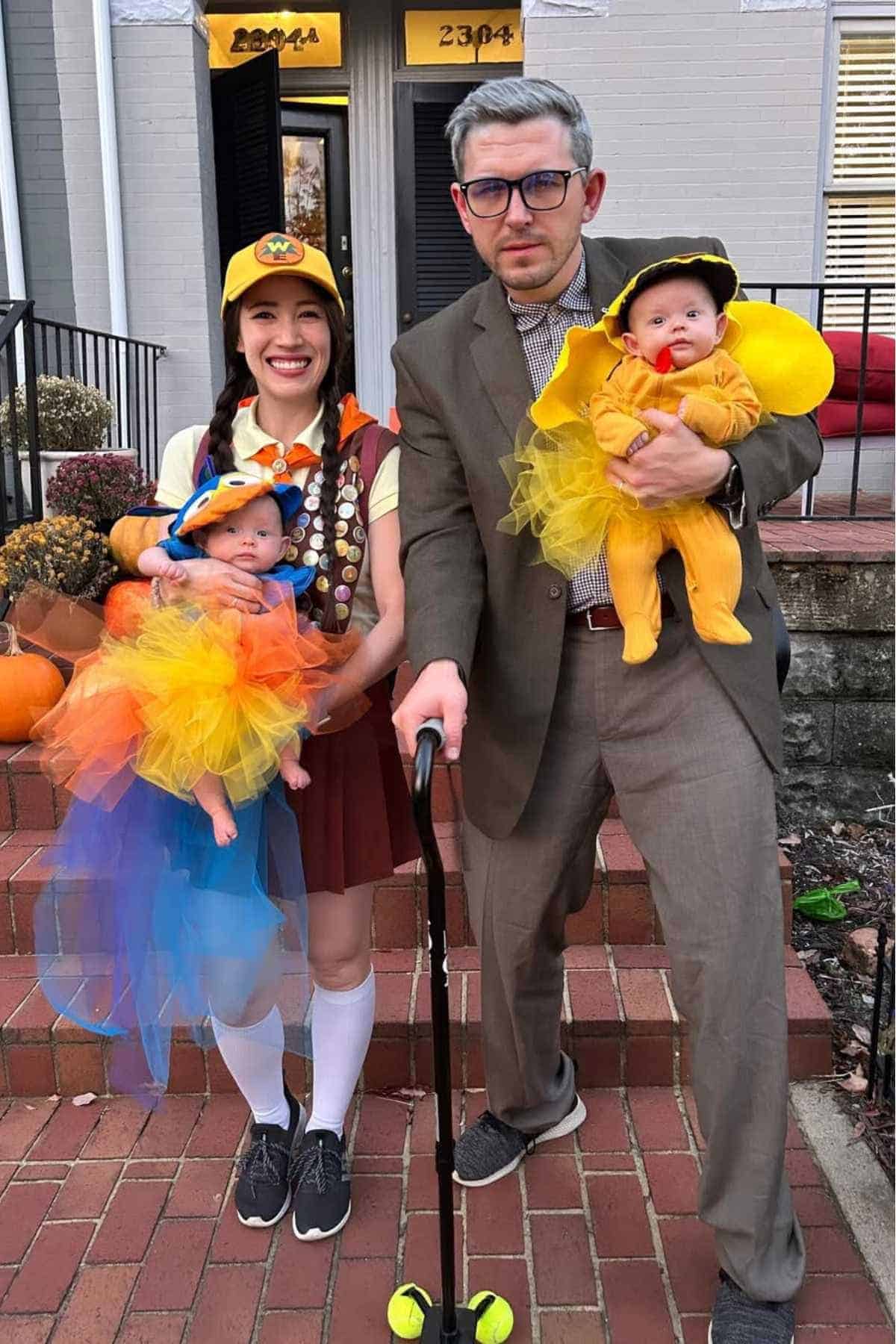 Two adults and two babies in costumes pose on steps; one adult is dressed as a scout, another in a suit with a cane, and the babies wear colorful tulle outfits.