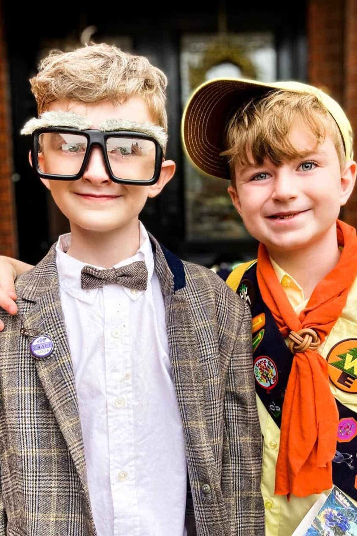 Two children dressed in costumes; one as an elderly man with large glasses and a bowtie, and the other as a scout with a yellow cap, orange scarf, and badges.