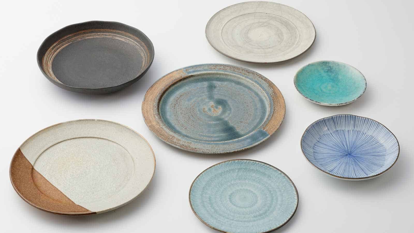 Seven ceramic plates of varying sizes and colors, including gray, blue, brown, and white, are arranged on a white surface.