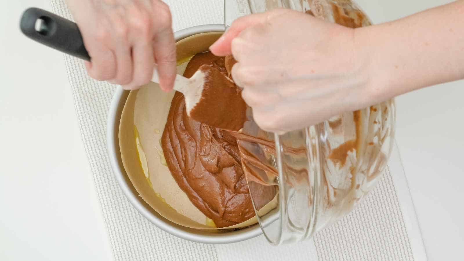 A person pours chocolate cake batter from a glass bowl into a round, parchment-lined baking pan using a spatula.