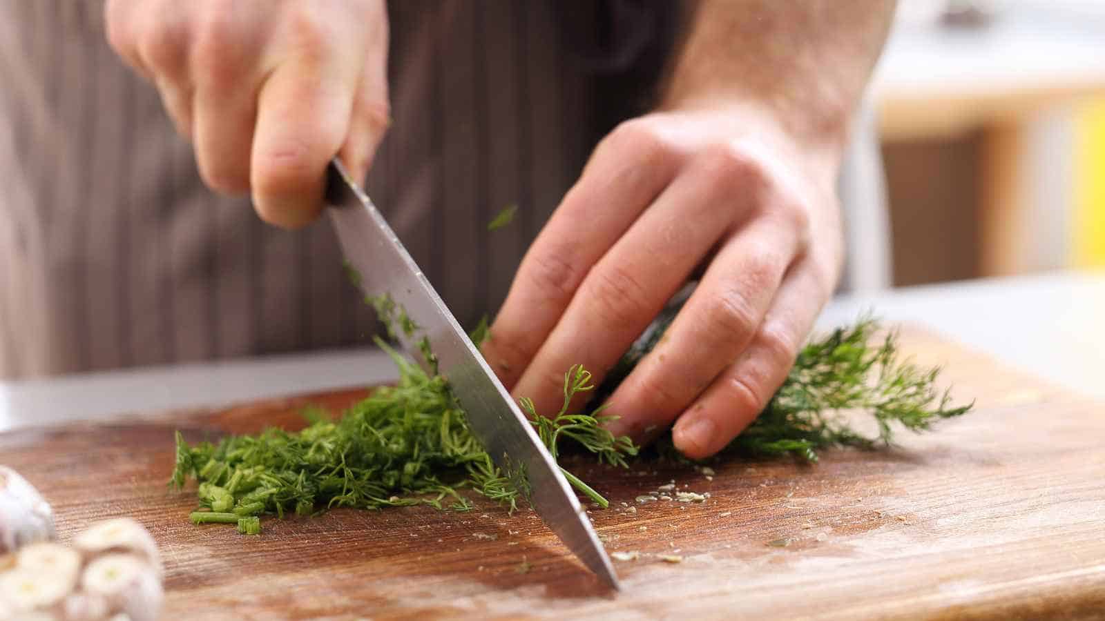 A person slices fresh herbs on a wooden cutting board with a large kitchen knife.