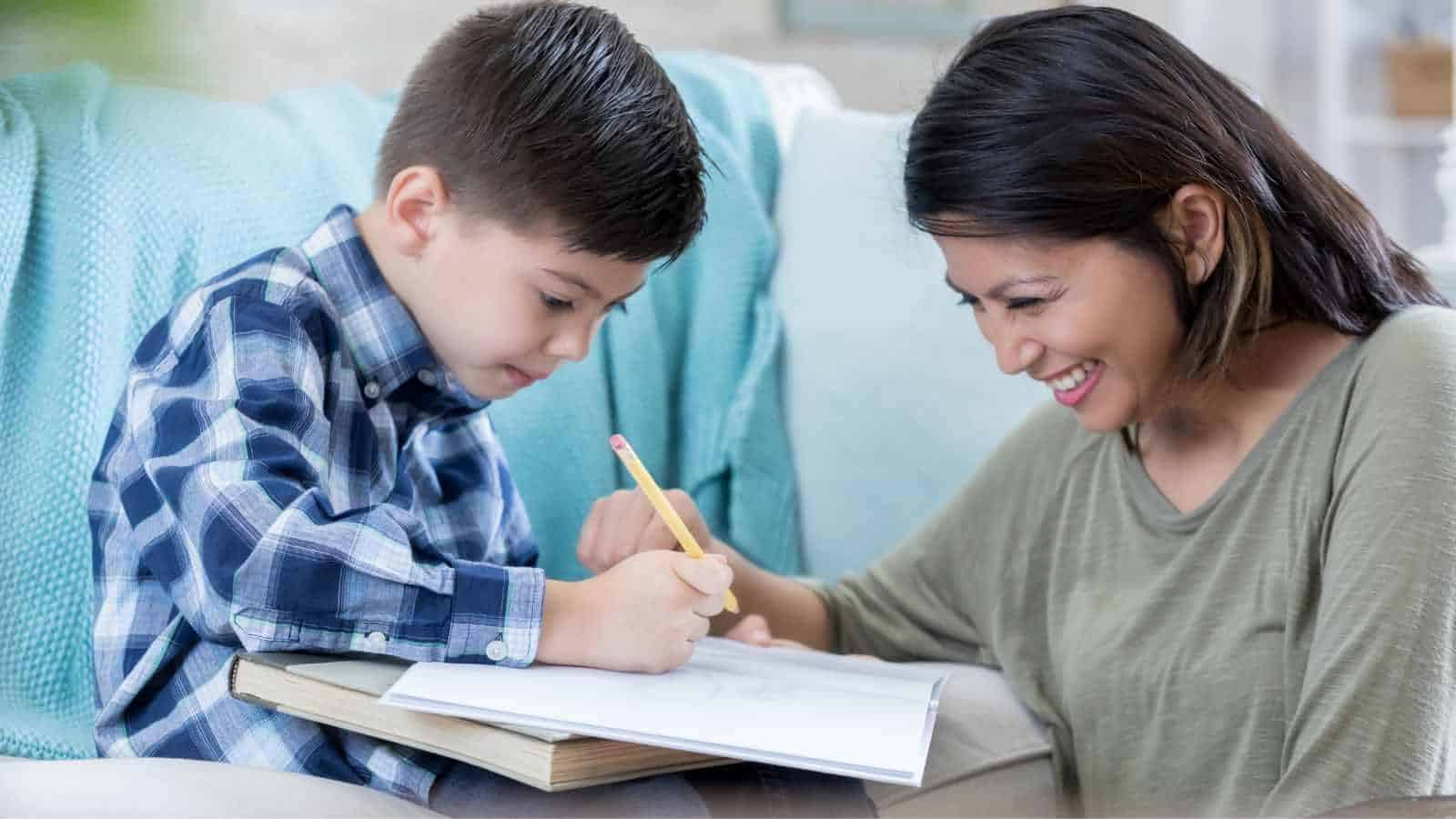 A young boy writes in a notebook with a pencil while an adult woman sits beside him, smiling and watching.
