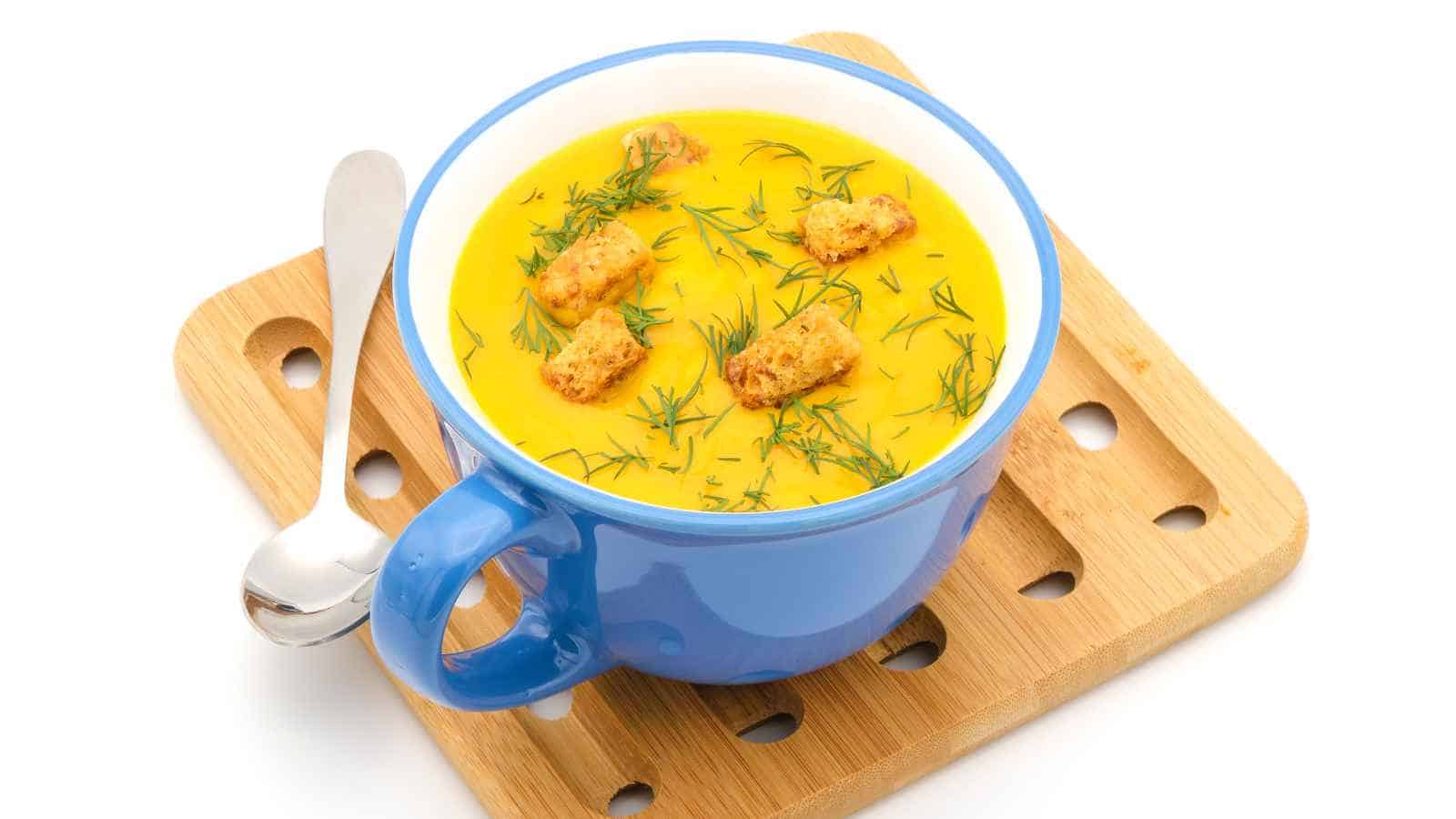 A blue cup filled with creamy yellow soup, garnished with croutons and fresh herbs, sits on a wooden tray with a metal spoon beside it.