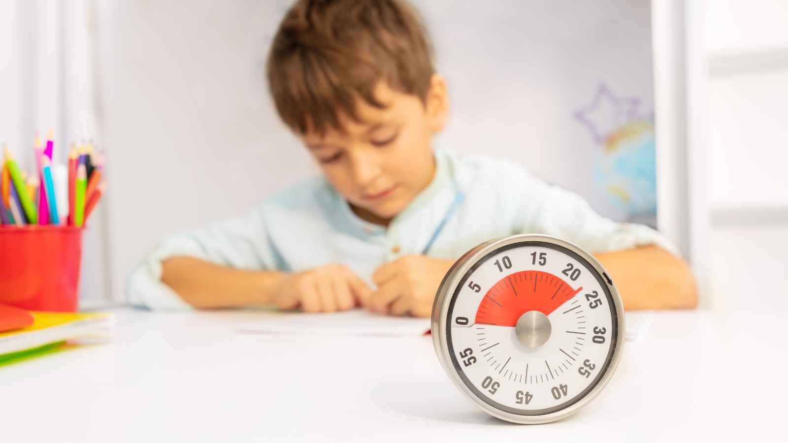 A young boy sits at a desk working on a task, with a timer set to 15 minutes placed in the foreground.