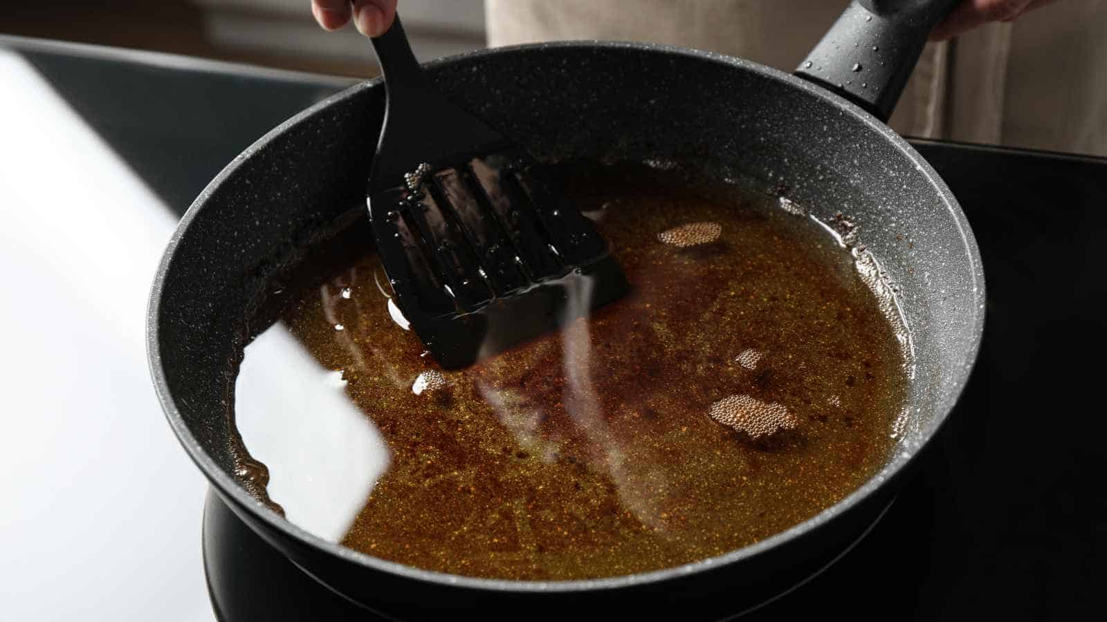 A hand holds a black spatula over a nonstick frying pan filled with hot oil on a stovetop.