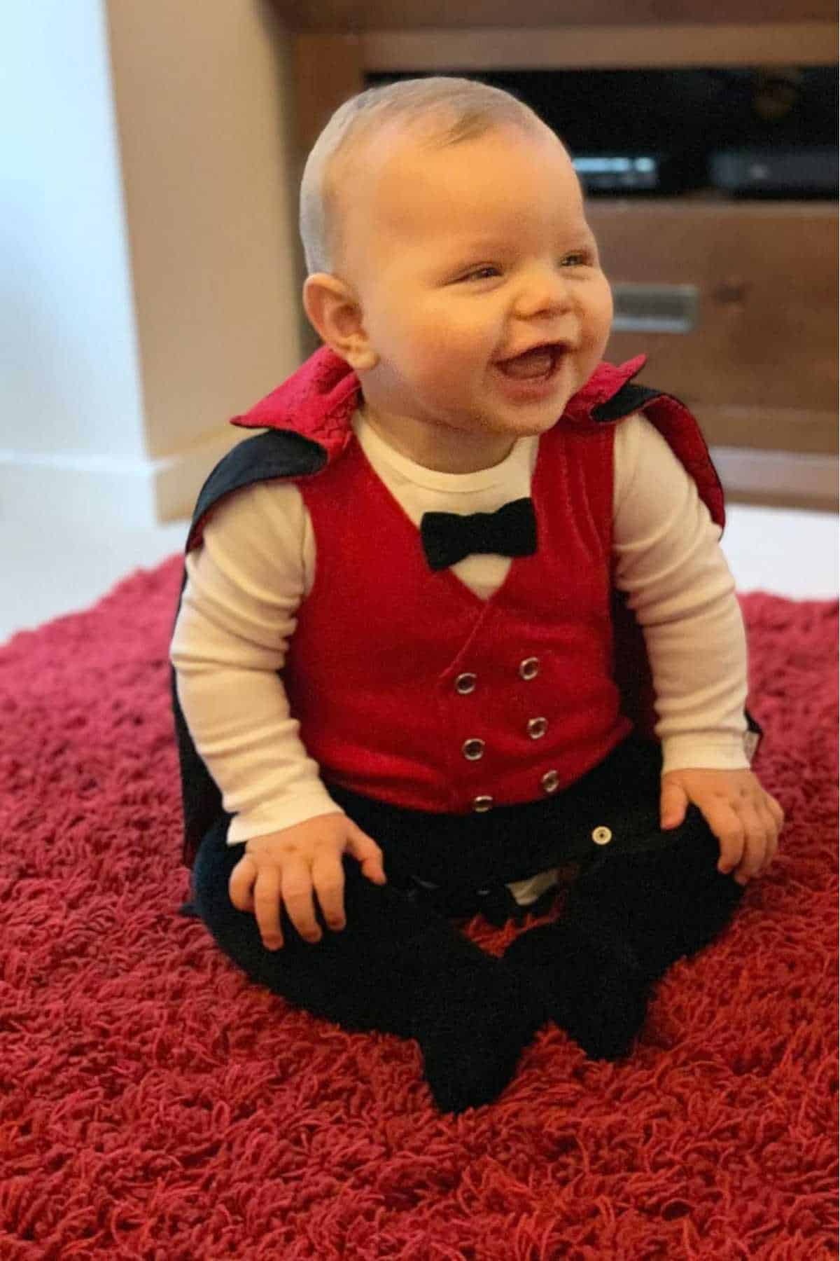 A smiling baby sits on a red carpet wearing a red vest, white shirt, black pants, black bow tie, and a black and red cape.