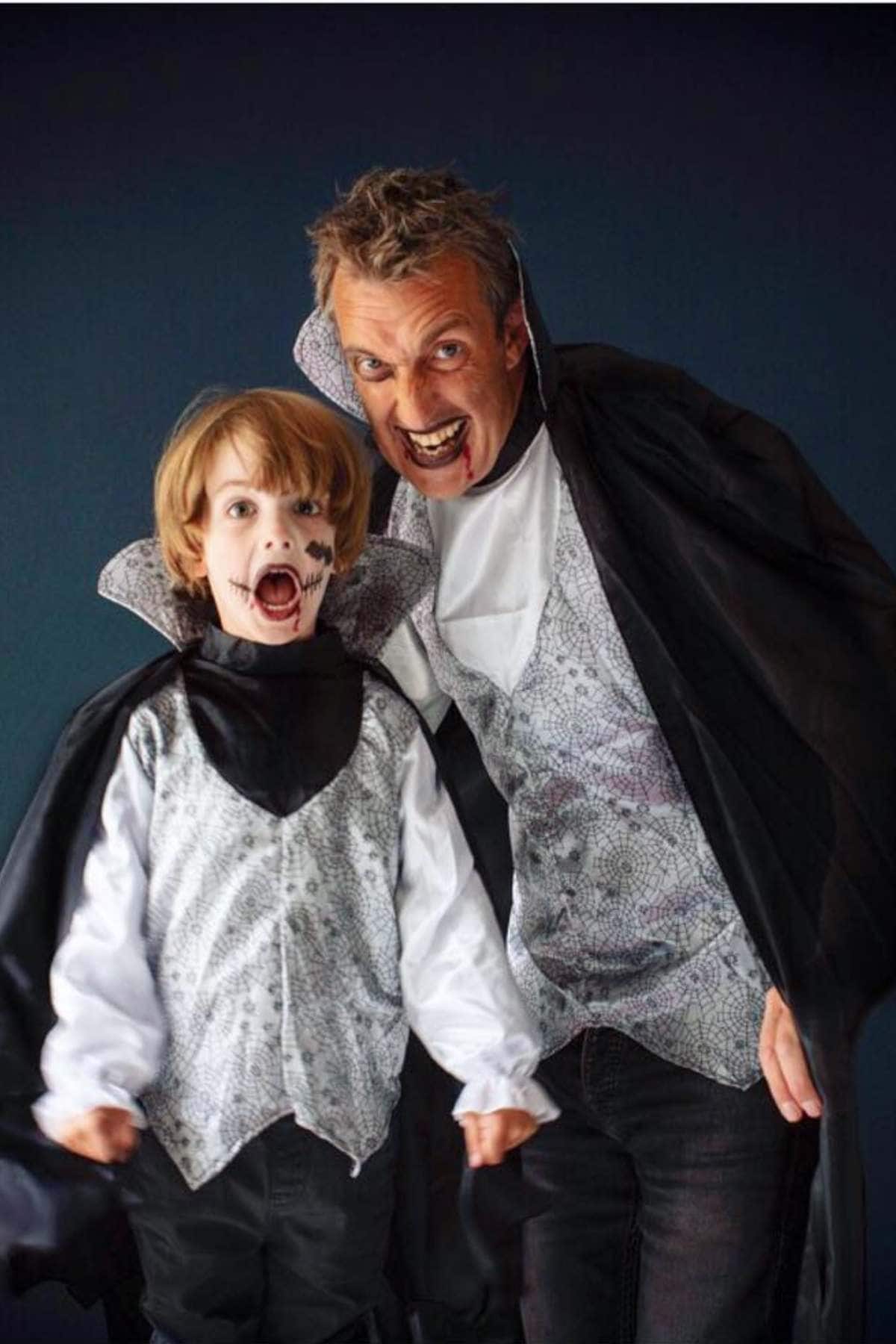 An adult and a child dressed in matching vampire costumes pose together, both with fangs bared and dramatic facial expressions.