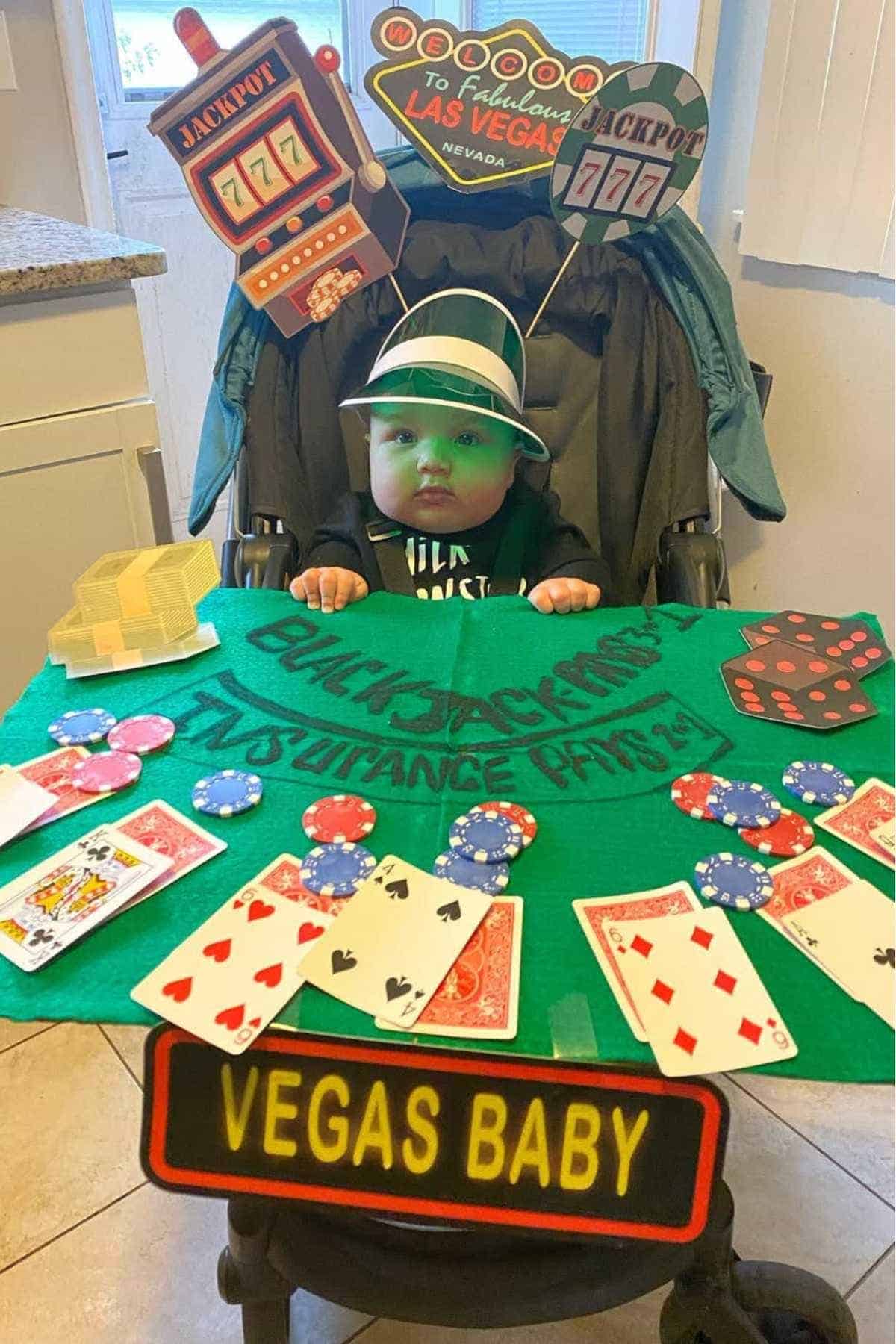 A baby in a stroller is dressed as a Vegas gambler, with a casino-themed display, poker chips, cards, and signs reading “Jackpot” and “Vegas Baby.”