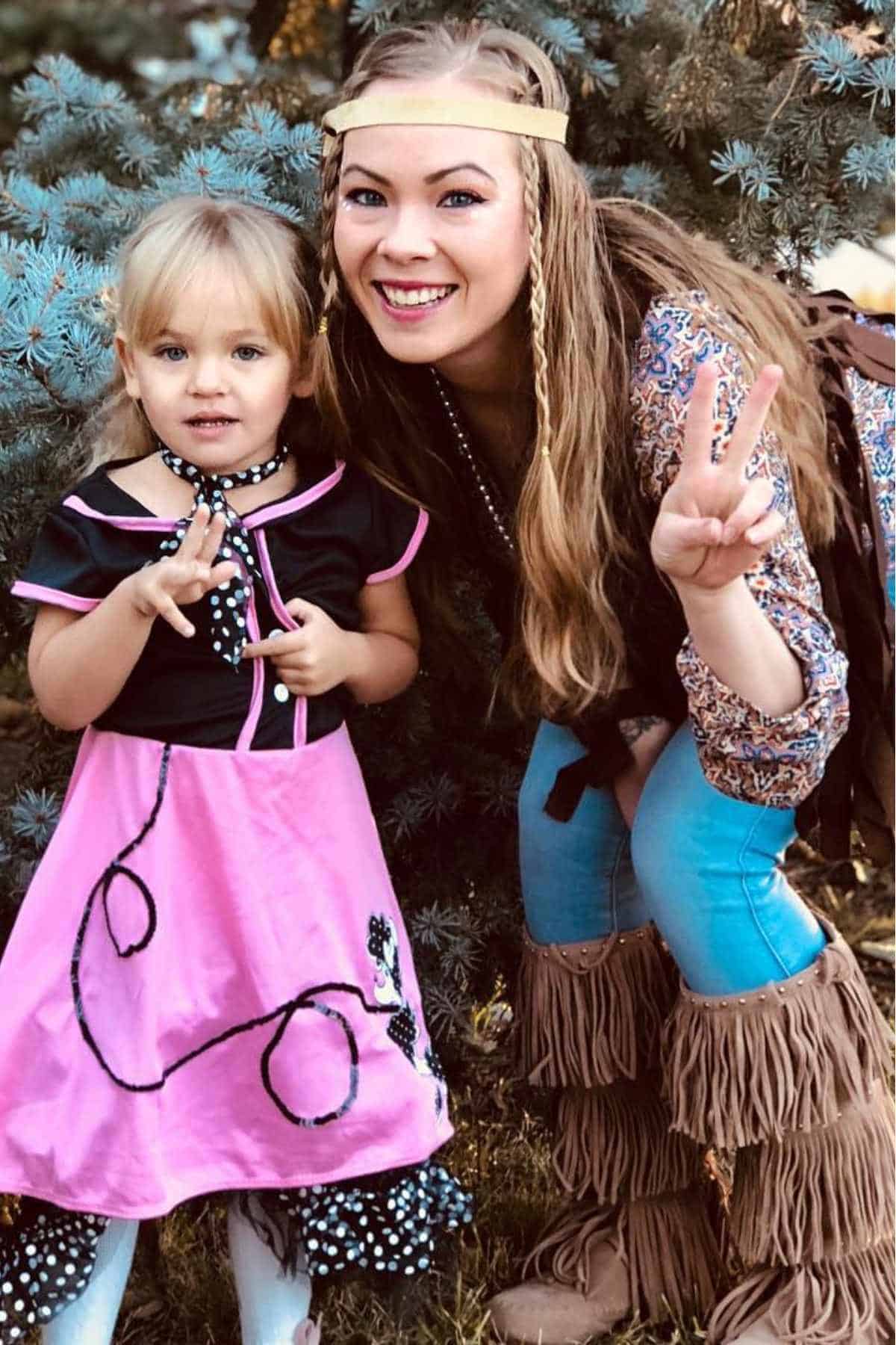 An adult and a child dressed in costumes pose outside; the child wears a pink poodle skirt dress, and the adult wears a patterned top, headband, and fringed boots.
