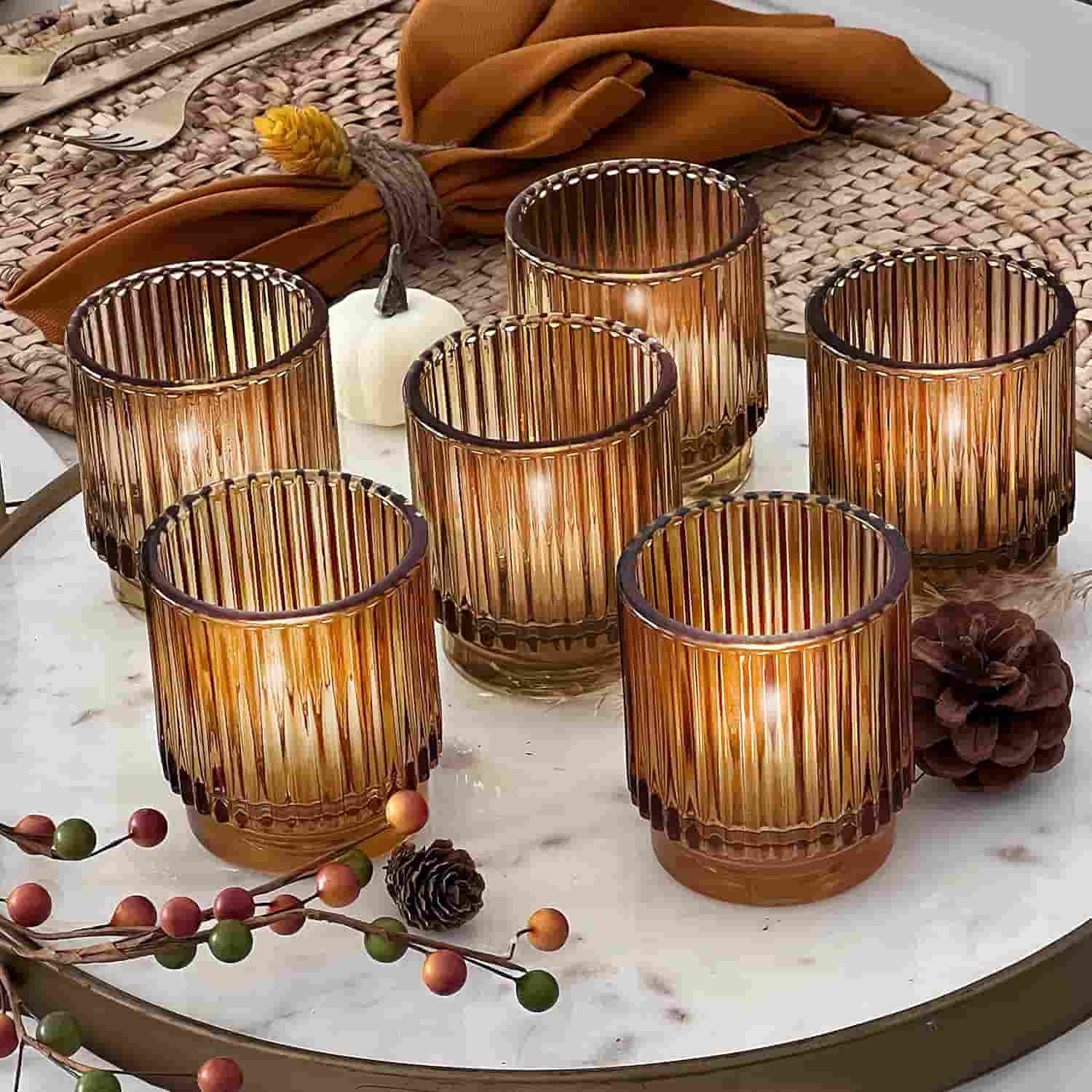 Six ribbed amber glass candle holders with lit candles are arranged on a marble tray, surrounded by autumn-themed decor, including pinecones and berries.