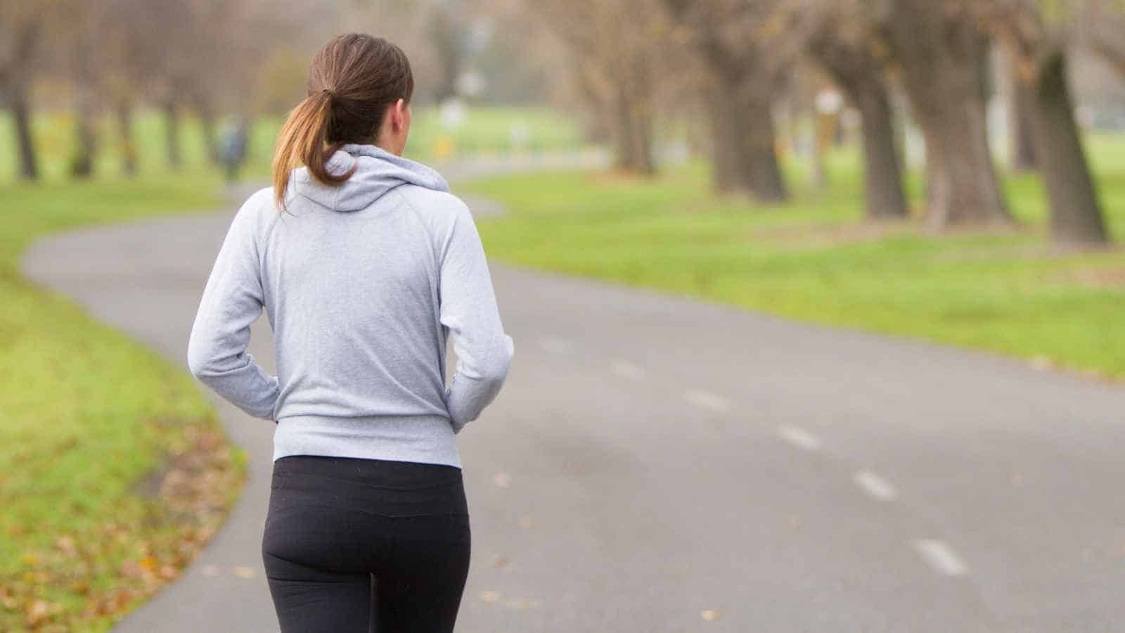 A person wearing a gray hoodie and black pants jogs along a paved path in a park with trees and grass on both sides.
