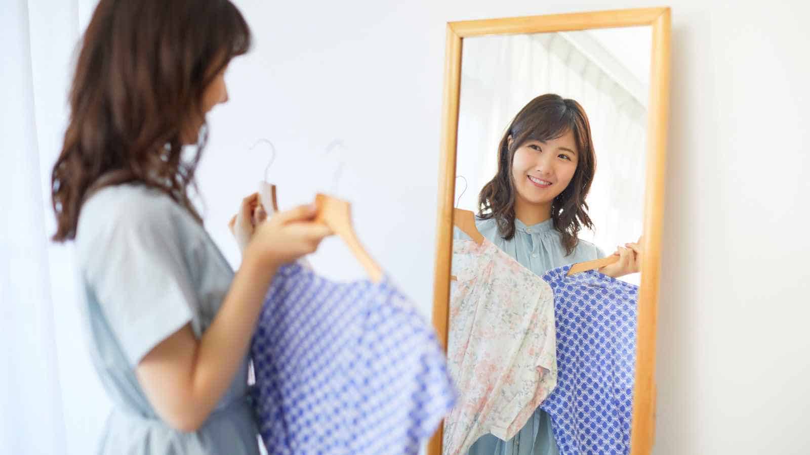 A woman stands in front of a mirror holding up two patterned shirts, appearing to decide which one to wear.