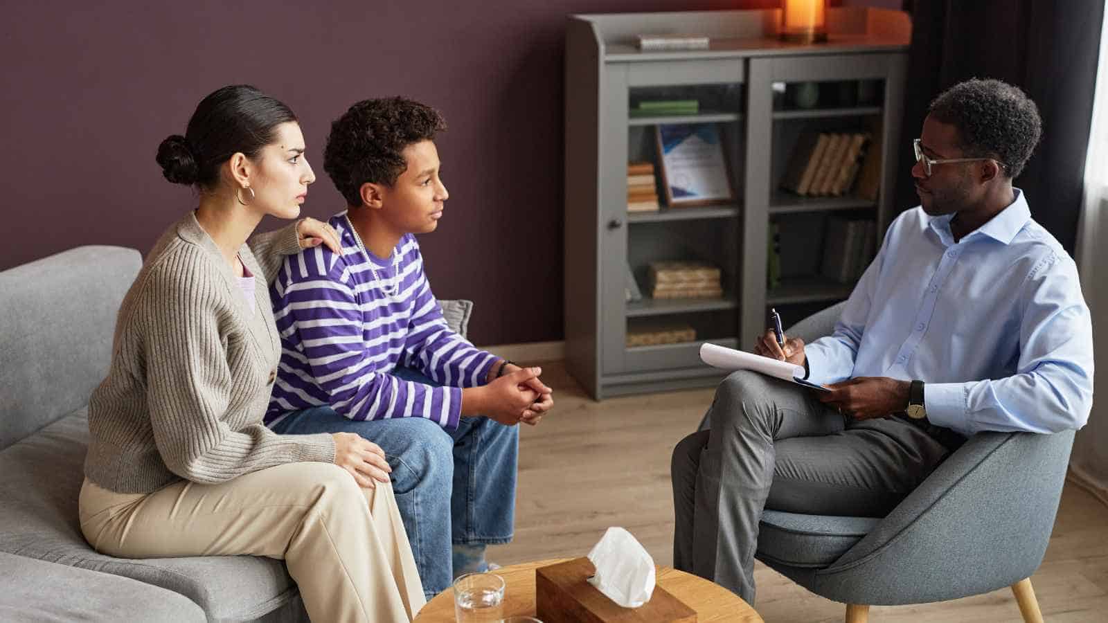 A woman and a boy sit on a couch talking to a man with a notebook in an office setting, suggesting a counseling or therapy session.