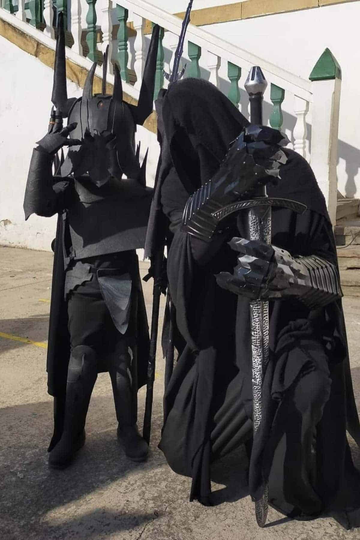 Two people dressed in detailed black costumes resembling Witch King standing outdoors near a staircase.